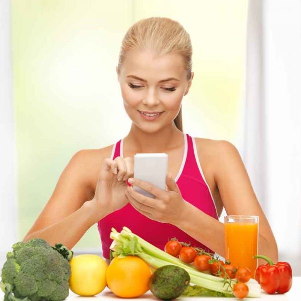 Fit woman sitting infront of fruits and veggies while counting macros using cellphone